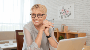 An older women smiling sitting at a desk with a laptop in front of her.